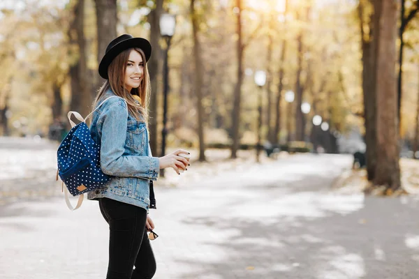Imagen de una chica hipster sonriente brillante con cabello castaño usando un sombrero, gafas de sol y mochila sosteniendo una taza de té o café en el parque — Foto de Stock