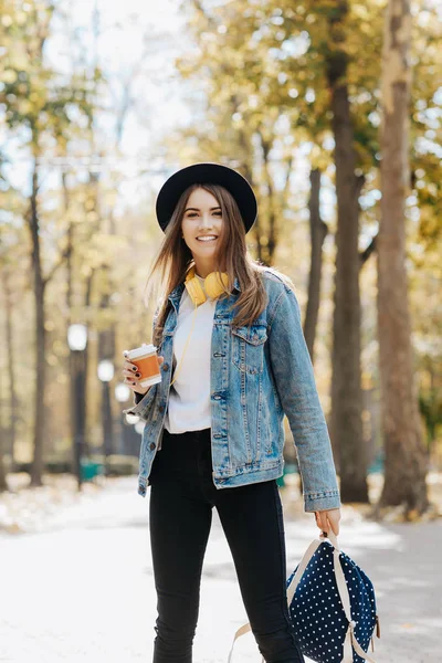 Image of a bright smiling hipster girl with brown hair wearing a hat, sunglasses and backpack holding cup of tea or coffee in the park