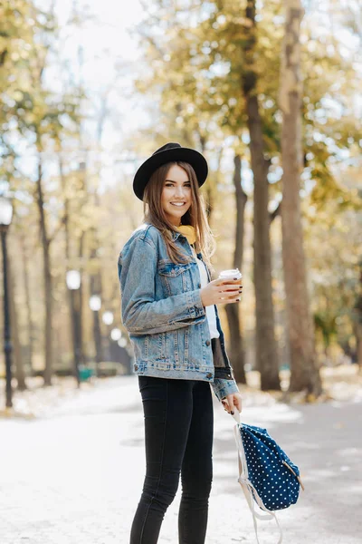 Image of a bright smiling hipster girl with brown hair wearing a hat, sunglasses and backpack holding cup of tea or coffee in the park