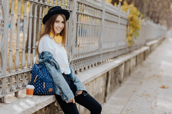 Photo of a bright smiling hipster girl with brown hair wearing a hat, sunglasses and backpack holding cup of tea or coffee in the park — Stock Photo, Image