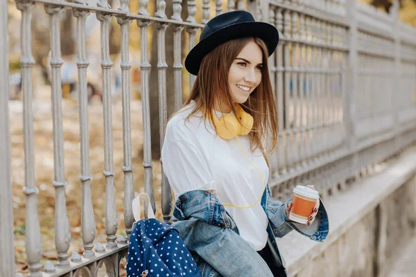 Close-up photo of a bright smiling hipster girl with brown hair wearing a hat, sunglasses and backpack holding cup of tea or coffee in the park — Stock Photo, Image