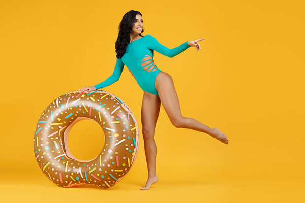 Full lenght portrait of an excited young fit woman dressed in swimsuit posing with inflatable ring and pointing finger away on the orange background.