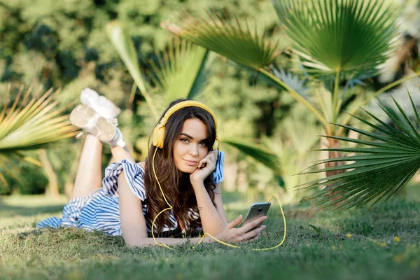 Ritratto di una bella ragazza in abiti estivi che ascolta musica con le cuffie gialle mentre tiene il cellulare sull'erba tra le palme. Concetto di libertà . — Foto Stock