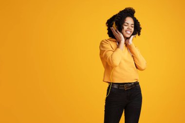 Trendy Afrikalı-Amerikalı genç kadın parlak bir gülümseme ile rahat kıyafetler ve kulaklık müzik dinleme uzakta sarı arka plan üzerinde giyim giyinmiş