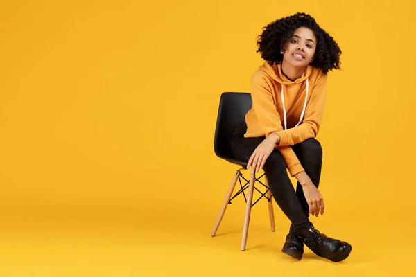 Porträtt av charmiga afroamerikanska ung kvinna med vackert leende klädd i casual kläder sitter på den eleganta stolen över gul bakgrund. — Stockfoto