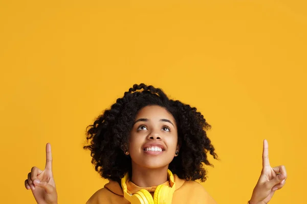 Charmante afrikanisch-amerikanische junge Frau mit strahlendem Lächeln in lässiger Kleidung und Kopfhörer-Kleidung, die Finger nach oben über gelbem Hintergrund zeigt. — Stockfoto