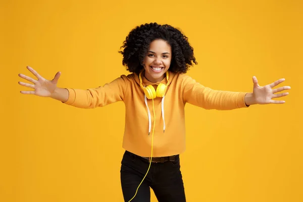 Schöne afrikanisch-amerikanische junge Frau mit strahlendem Lächeln in lässiger Kleidung und Kopfhörer bereit für Umarmungen vor gelbem Hintergrund. — Stockfoto