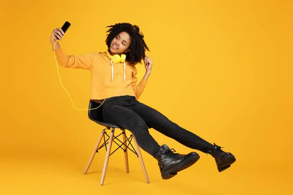 Porträt einer lustigen afrikanisch-amerikanischen jungen Frau mit schönem Lächeln in lässiger Kleidung und Kopfhörern, die auf dem Stuhl sitzt und ein Selfie mit dem Smartphone vor gelbem Hintergrund macht. — Stockfoto