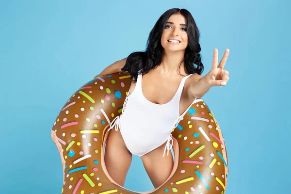 Retrato de una joven excitada vestida con traje de baño blanco posando con anillo inflable y mostrando gesto de paz sobre el fondo azul . — Foto de Stock