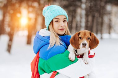 Portrait of cheerful young woman in warm stylish clothes and bagpack posing with her beagle dog in winter park. Friendship, pet and human. clipart
