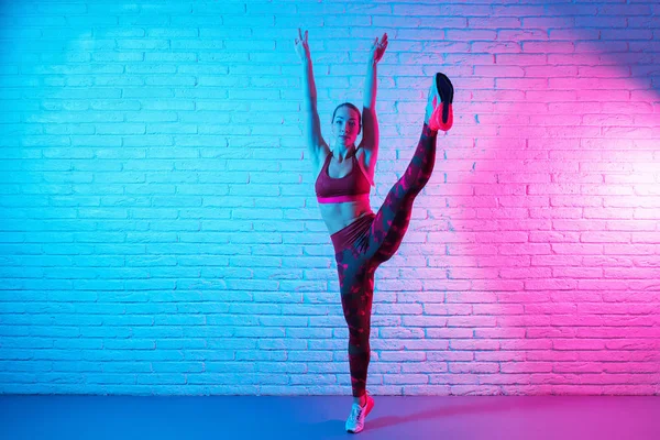 Hübsche junge schlanke Turnerin in Sportkleidung, die sich vor einer Ziegelwand im Neonlicht ausbreitet. Flexible muskulöse Frau turnt Spagat. — Stockfoto