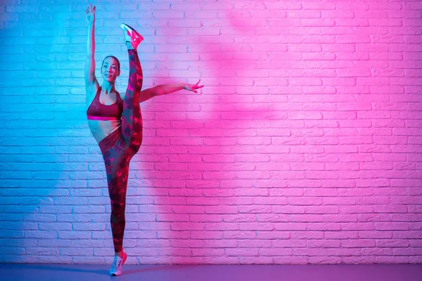 Charmante junge schlanke Turnerin in Sportkleidung, die sich vor einer Ziegelwand im Neonlicht ausbreitet. Flexible muskulöse Frau turnt Spagat. — Stockfoto