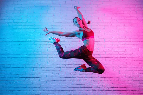 Charmante junge schlanke Turnerin in Sportkleidung turnt vor einer Ziegelwand im Neonlicht. Flexible muskulöse Frau turnt Elemente. — Stockfoto