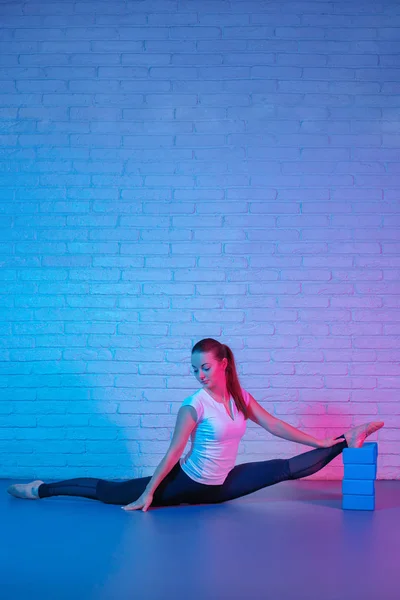 Hübsche junge, schlanke Turnerin in Sportkleidung, die sich im Neonlicht gegen eine Ziegelwand reckt. Flexible muskulöse Frau macht fortgeschrittenen gymnastischen Spagat. — Stockfoto