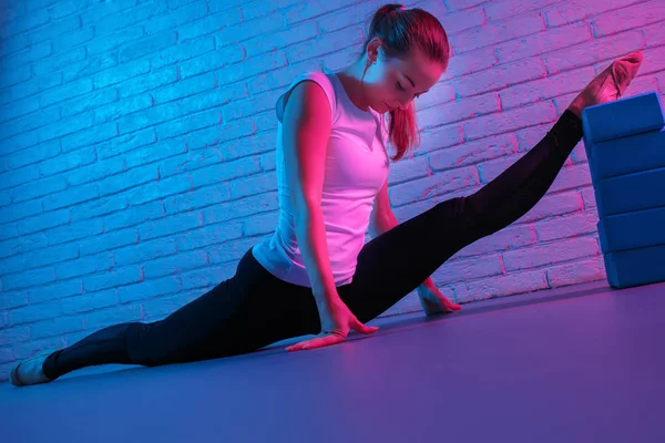 Neon ışıkları tuğla duvara germe giyim spor yakışıklı genç ince jimnastikçi kadın. Esnek kaslı kadının yapması jimnastik split gelişmiş. — Stok fotoğraf