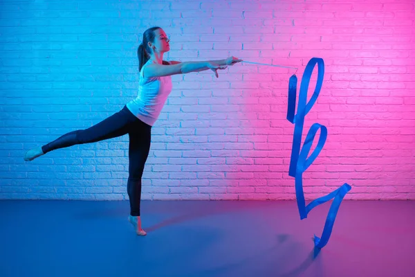 Hübsche junge schlanke Turnerin in Sportkleidung, die sich vor einer Ziegelwand im Neonlicht ausbreitet. flexible muskulöse Frau macht Spiralen mit Schleife. — Stockfoto