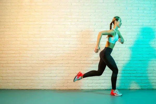 Sportif, genç ve çekici bir kadın. Neon ışıklarıyla ön tuğla duvarda koşuyor.. — Stok fotoğraf