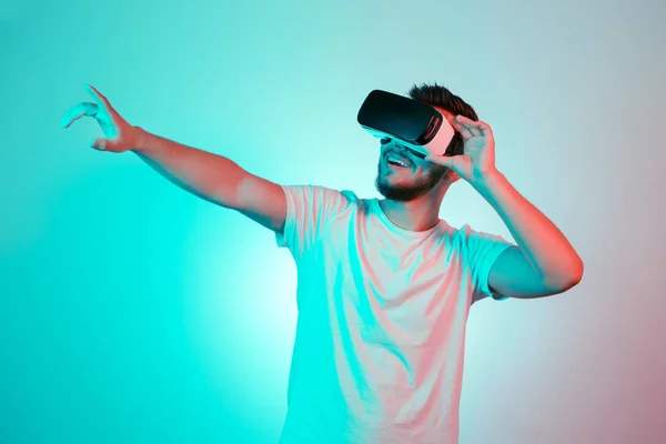 Attractive bearded man trying VR headset and poniting. Young man exploring another world with virtual reality goggles on the colorful background.