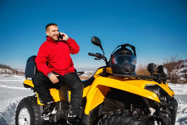 Attractive young man driver in red warm winter clothes and black helmet talking on smartphone near the ATV 4wd quad bike stand in heavy snow with deep wheel track. Moto winter sports.