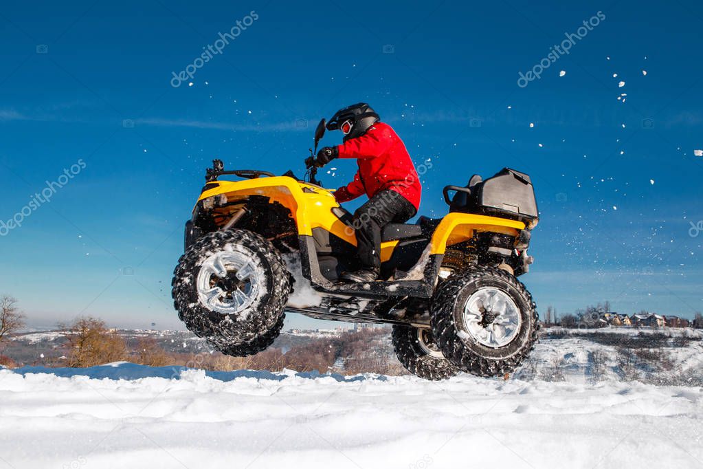 Photo of young extremal driver in red warm winter clothes and black helmet jump in the air with the ATV 4wd quad bike stand in heavy snow with deep wheel track. Extreme moto winter sports