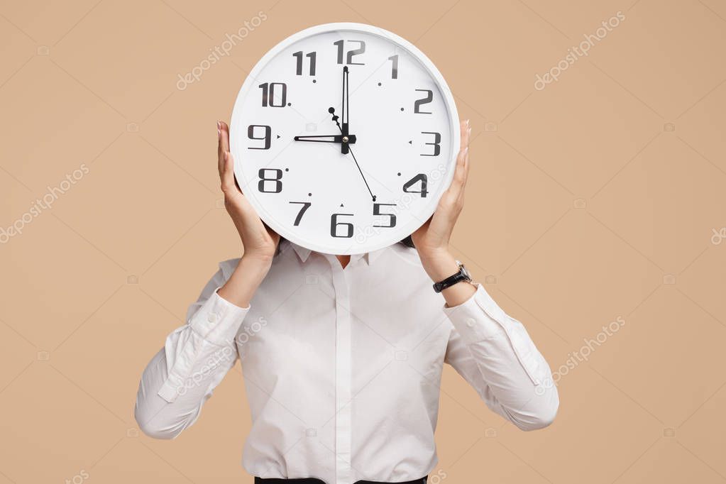 Photo of young modern business woman hold a clock in front of the face over beige background.