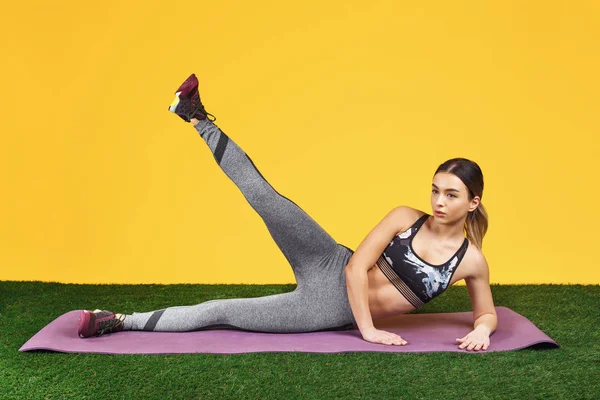Довольно подходящая молодая женщина делает упражнения на фитнес-фиолетовый мат на зеленой траве на желтом фоне . — стоковое фото