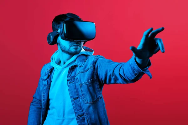 Çekici sakallı adam VR kulaklık çalışıyor ve Neon ışıkta kırmızı arka planda stüdyoda başka bir dünya keşfetmek. VR kulaklık ile kullanarak Smartphone.