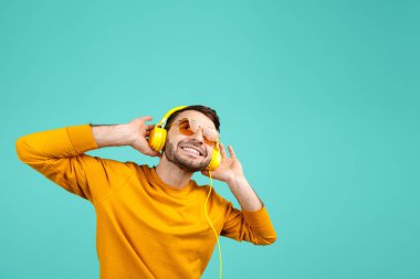 Sarı güneş gözlüğü giyen yakışıklı sakallı genç adam mavi arka planda sarı kulaklıklar ile müzik dinlerken.