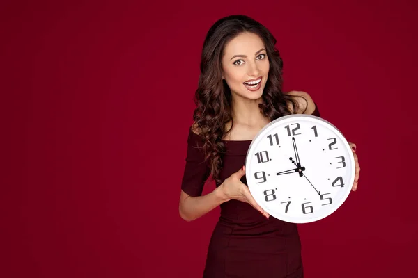 Foto von hellen jungen modernen Geschäftsfrau in kastanienbraunem Kleid hält eine Uhr über rotem Hintergrund, — Stockfoto