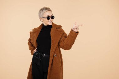 Mutlu genç hipster kadın sarışın kısa saçlı bir ceket ve güneş gözlüğü noktası parmak uzakta bej renkli arka plan üzerinde izole giyiyor.