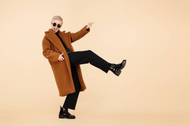 Çekici genç hipster kadın sarışın kısa saçlı bir ceket ve güneş gözlüğü üzerinde görünmez gitar çalmak giyen ile bej renkli arka plan üzerinde izole. Tam lengh fotoğraf.