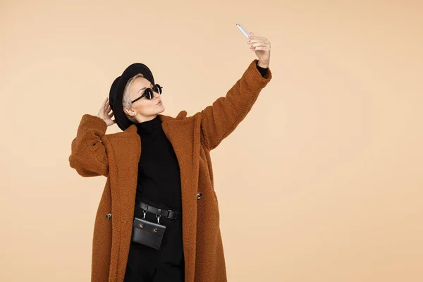 Pretty young hipster woman with blonde short hair wearing a coat, hat and sunglasses make selfie with smartphone isolated over beige background. — Stock Photo, Image