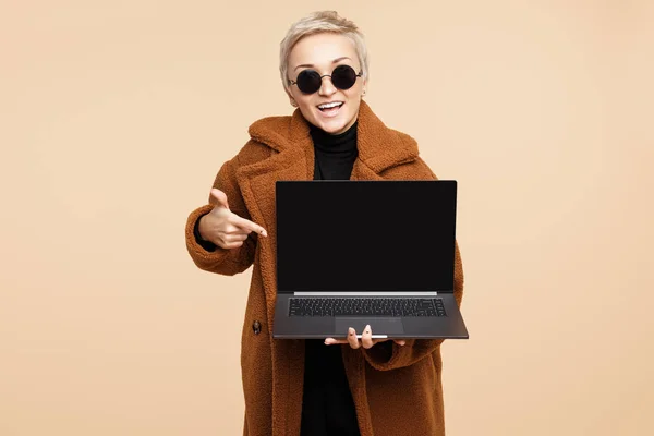 Shocked young hipster woman with blonde short hair wearing a coat and sunglasses point finger on the computer or laptop isolated over beige background. — Stock Photo, Image
