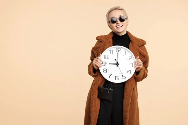Glad ung hipster kvinna med blont kort hår iklädd en kappa och solglasögon håller en klocka som isolerade över beige bakgrund. — Stockfoto