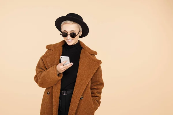 Trendy young hipster woman with blonde short hair wearing a coat, hat and sunglasses use smartphone isolated over beige background. — Stock Photo, Image