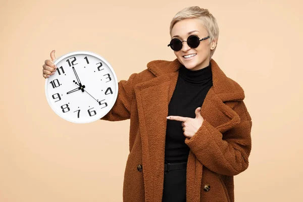 Glückliche junge Hipsterfrau mit blonden kurzen Haaren, die einen Mantel und eine Sonnenbrille trägt, pint Finger auf der Uhr isoliert über beigem Hintergrund. — Stockfoto