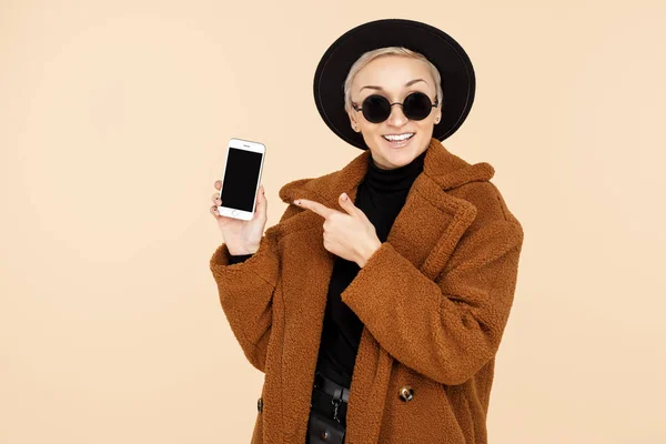 Charming young hipster woman with blonde short hair wearing a coat, hat and sunglasses point finger on the smartphone isolated over beige background. — Stock Photo, Image