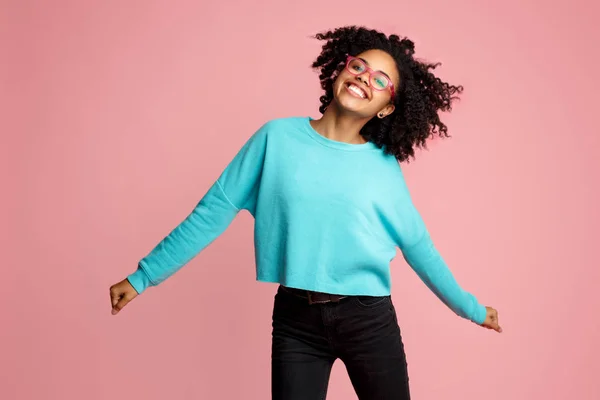 Upphetsad afroamerikanska ung kvinna med ljusa leende klädd i casual kläder, Glasögon och hörlurar Dans över rosa bakgrund. — Stockfoto