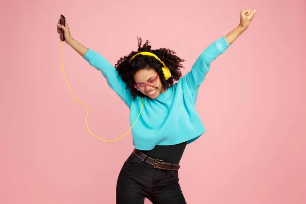 Ganska afroamerikanska ung kvinna med ljusa leende klädd i casual kläder, glasögon och hörlurar dans över rosa bakgrund. — Stockfoto