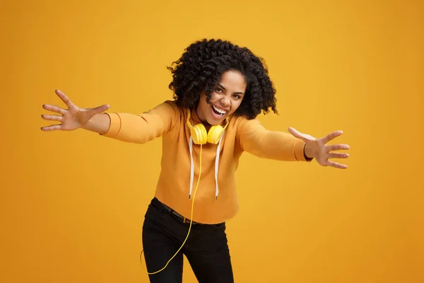 Schöne afrikanisch-amerikanische junge Frau mit strahlendem Lächeln in lässiger Kleidung und Kopfhörer bereit für Umarmungen vor gelbem Hintergrund. — Stockfoto
