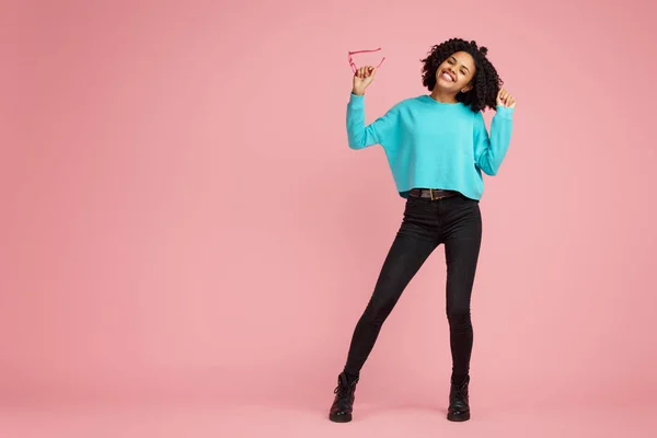 Full lengh foto av glada afrikanska amerikanska ung kvinna med ljusa leende klädd i casual kläder, glasögon och hörlurar dans över rosa bakgrund. — Stockfoto