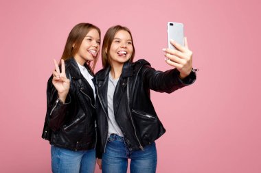 Parlak gülümseme ile deri ceketleri muhteşem iki genç ikiz kız pembe arka plan üzerinde akıllı telefon kullanın. İkiz kardeşler cep telefonu ile selfie almak.