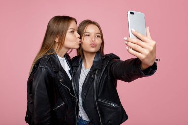 Pembe arka plan üzerinde parlak gülümseme kullanımı akıllı telefon ile deri ceketler güzel iki genç ikiz kız kardeş. İkiz kardeşler cep telefonu ile selfie almak.