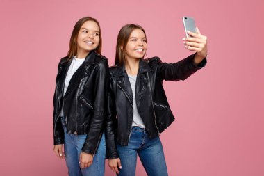 Pembe arka plan üzerinde parlak gülümseme kullanımı akıllı telefon ile deri ceketler güzel iki genç ikiz kız kardeş. İkiz kardeşler cep telefonu ile selfie almak.