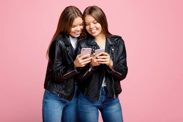 Прекрасні дві молоді сестри-близнючки з яскравою посмішкою в шкіряних куртках використовують смартфон на рожевому фоні . — стокове фото