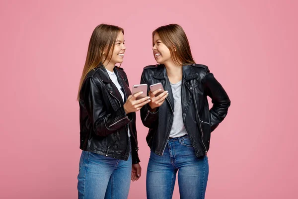 Прекрасні дві молоді сестри-близнючки з яскравою посмішкою в шкіряних куртках використовують смартфон на рожевому фоні . — стокове фото