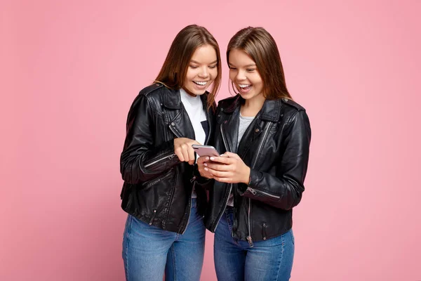 Funny two young twin sisters with bright smile in leather jackets use smartphone over pink background. — Stock Photo, Image