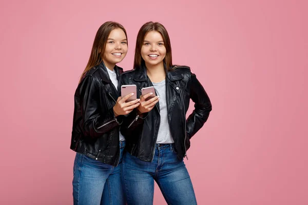 Хороші дві молоді сестри-близнючки з яскравою посмішкою в шкіряних куртках використовують смартфон на рожевому фоні . — стокове фото
