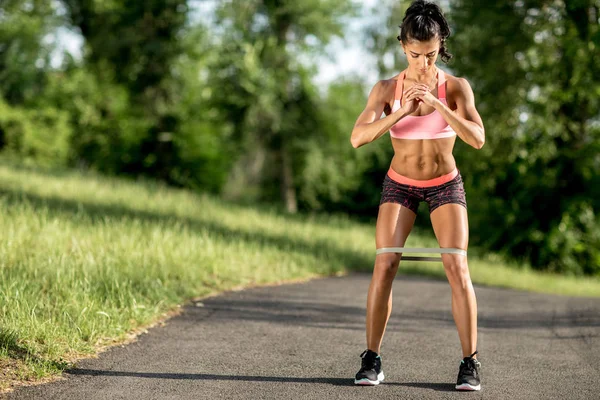 Jovem mulher esportiva fazendo exercícios de agachamento com elástico ao ar livre . — Fotografia de Stock