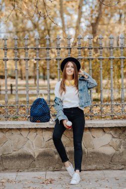 Parkta şapka, sırt çantası ve sarı kulaklık takan kahverengi saçlı güzel, gülümseyen hipster kız öğrenciportresi.